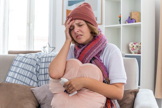 Unhealthy young woman in warm hat with scarf looking unwell holding pillow and tissue suffering from cold and flu having strong headache sitting on the chair in light living room