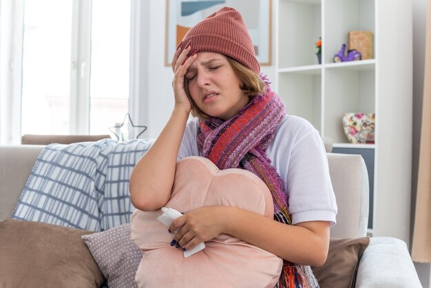 Нездоровая молодая женщина в теплой шляпе с шарфом, которая выглядит нездоровой, держит подушку и салфетку, страдает от простуды и гриппа и страдает от сильной головной боли, сидя на стуле в светлой гостиной