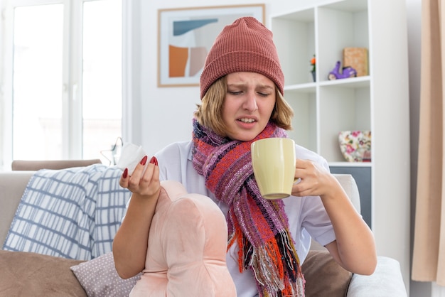 Unhealthy young woman in warm hat with scarf looking unwell and sick holding cup of hot tea and tissue suffering from cold and flu sitting on the chair in light living room