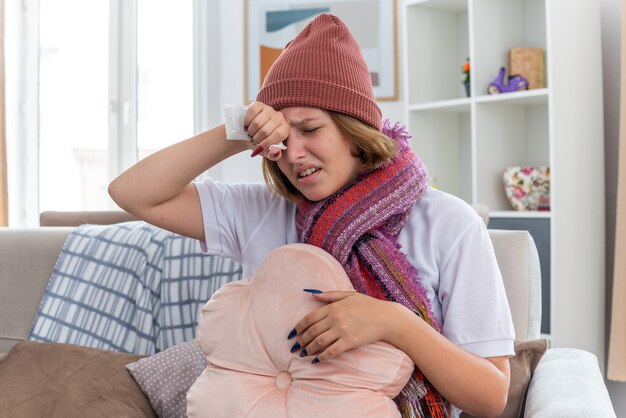 暖かい帽子をかぶった不健康な若い女性で、スカーフが体調が悪く、額に触れ、明るいリビング ルームのソファに座って、風邪やインフルエンザに苦しんでいる発熱と頭痛を抱えている