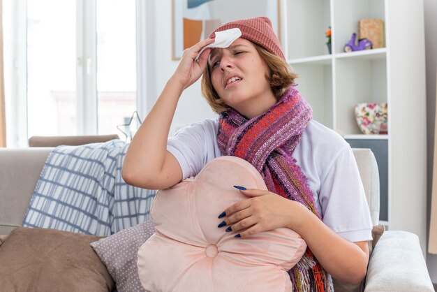 Unhealthy young woman in warm hat with scarf looking unwell and sick touching her forehead having fever and headache suffering from cold and flu sitting on couch in light living room