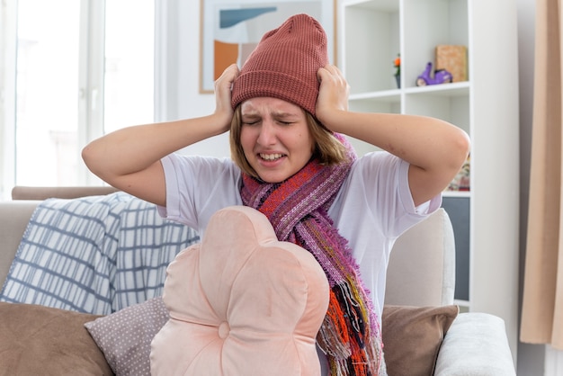 Unhealthy young woman in warm hat with scarf looking unwell and sick touching her head having fever and headache suffering from cold and flu sitting on couch in light living room