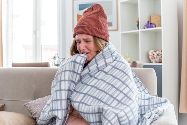 Unhealthy young woman in warm hat wrapped in blanket looking unwell and sick suffering from cold and flu having fever and headache sitting on couch in light living room