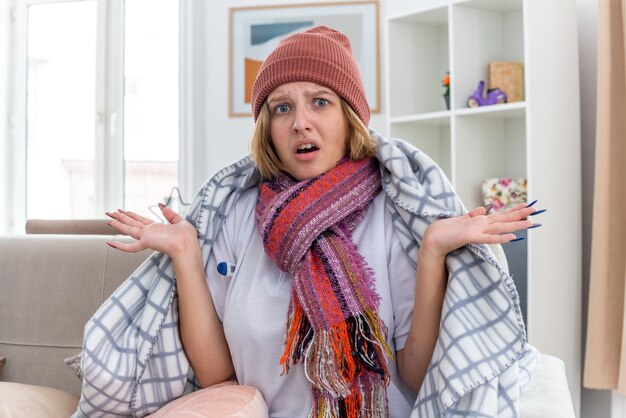 Unhealthy young woman in warm hat wrapped in blanket looking unwell and sick suffering from cold and flu with thermometer having fever looking confused sitting on couch in light living room