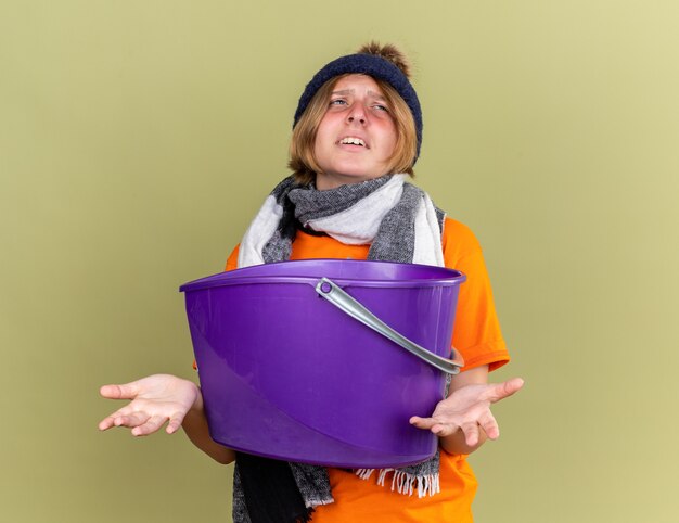 Unhealthy young woman wearing hat with scarf around her neck feeling sick suffering from nauseas holding basin looking confused standing over green wall