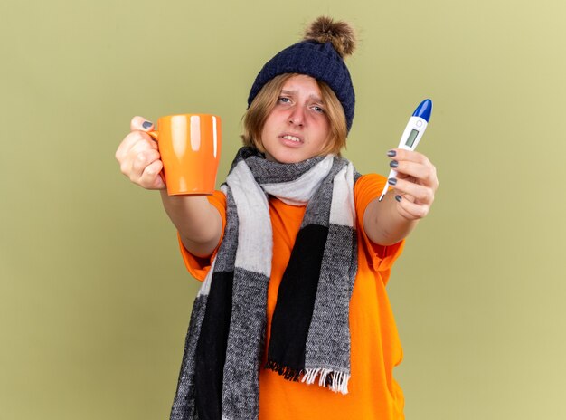 首にスカーフを巻いた帽子をかぶった不健康な若い女性が、インフルエンザと緑の壁の上に立つ熱に苦しんでいるデジタル体温計を持って熱いお茶を飲んでいる