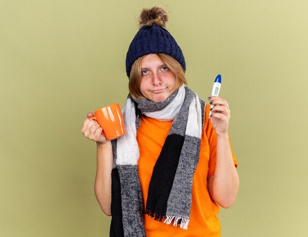 Нездоровая молодая женщина в шляпе с шарфом на шее чувствует недомогание, пьет горячий чай с цифровым термометром и страдает от гриппа и лихорадки
