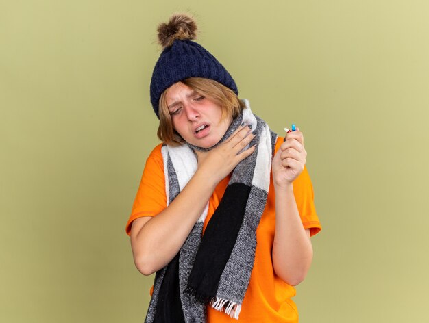インフルエンザや喉の痛みに苦しんでいるさまざまな錠剤を持ち、緑の壁の上に立って首に触れ、首にスカーフを巻いた帽子をかぶった不健康な若い女性