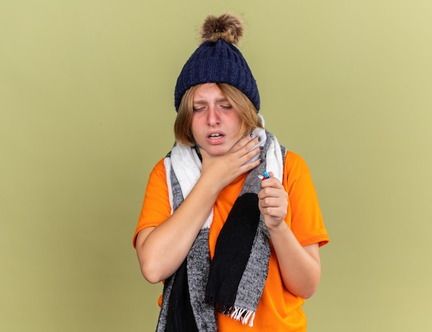 Unhealthy young woman wearing hat with scarf around her neck feeling unwell holding different pills suffering from throat sore touching feeling pain neck standing over green wall