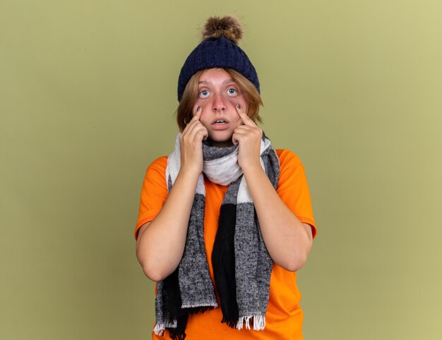 Unhealthy young woman wearing hat with scarf around her neck feeling unwell looking scared suffering from virus