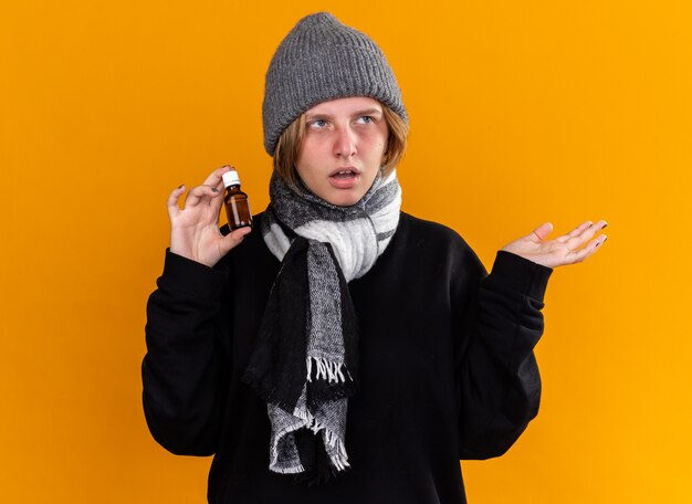 Unhealthy young woman wearing warm hat and with scarf around her neck feeling sick suffering from cold and flu holding medicine bottle presenting with arm standing over orange wall