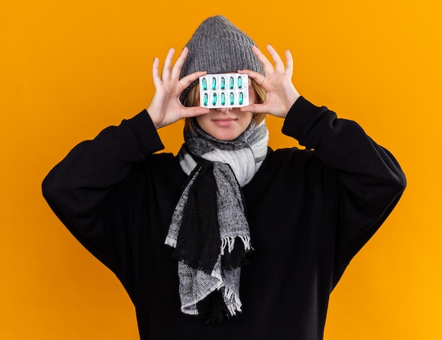 Foto gratuita giovane donna malsana che indossa un cappello caldo e con una sciarpa intorno al collo si sente malata soffre di raffreddore e influenza tenendo pillole sugli occhi