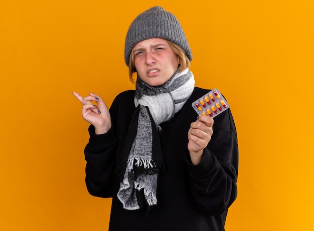 Unhealthy young woman wearing warm hat and with scarf around her neck feeling sick suffering from cold and flu holding pills poiting with index finger to the side standing over orange wall