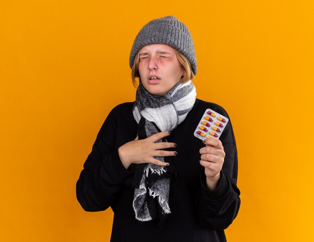 Нездоровая молодая женщина в теплой шляпе и с шарфом на шее, чувствуя себя больной от простуды и гриппа, держит таблетки, стоя над оранжевой стеной
