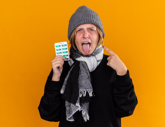 Unhealthy young woman wearing warm hat and with scarf around her neck feeling sick suffering from cold and flu holding pills sticking out tongue pointing with index finger on orange wall