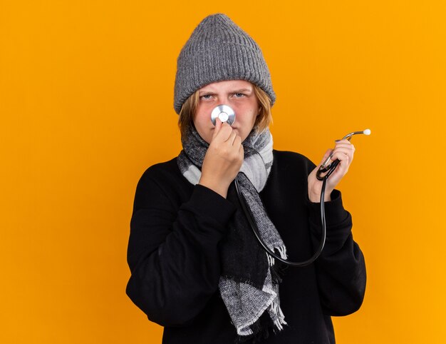 Unhealthy young woman wearing warm hat and with scarf around her neck feeling sick suffering from cold and flu holding stethoscope