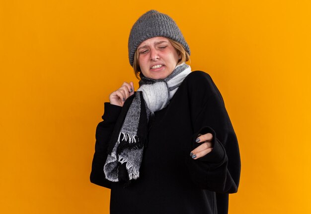 Нездоровая молодая женщина в теплой шапке и с шарфом на шее чувствует себя больной, страдает от простуды и гриппа, смотрит в сторону с отвращением