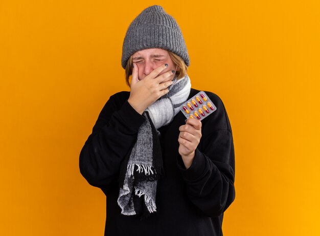Unhealthy young woman wearing warm hat and with scarf around her neck feeling terrible suffering from cold and flu holding pills covering mouth with hand sneezing standing over orange wall