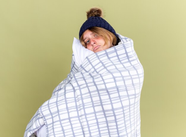 Unhealthy young woman wearing warm hat wrapped with blanket holding pillow feeling unwell suffering from cold standing over green wall