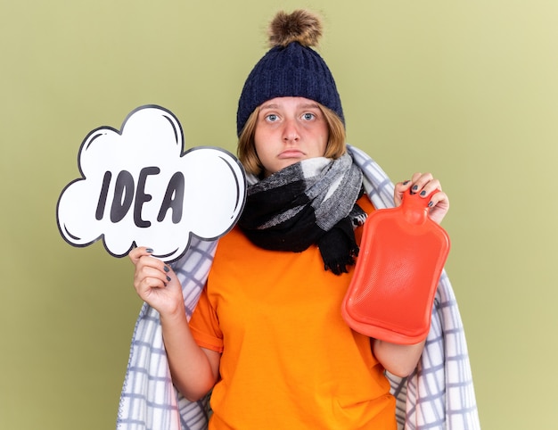 Free photo unhealthy young woman with warm hat and scarf around neck wrapped in blanket feeling unwell caught cold holding hot-water bottle and speech bubble sign with word idea
