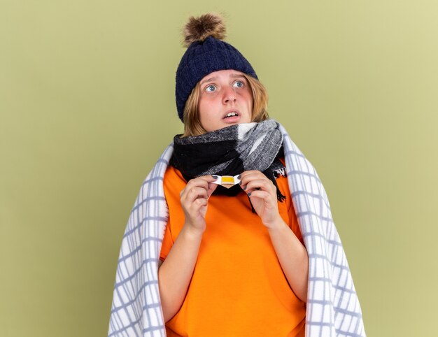 Unhealthy young woman with warm hat and scarf around neck wrapped in blanket feeling unwell holding a patch looking confused and worried