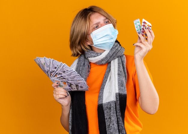 Нездоровая молодая женщина с теплым шарфом на шее в защитной маске для лица, держащая таблетки и деньги, выглядит смущенной