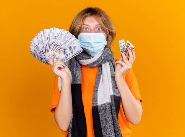 Unhealthy young woman with warm scarf around her neck wearing protective facial mask holding pills and cash looking surprised and confused standing over orange wall