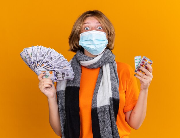 Нездоровая молодая женщина с теплым шарфом на шее в защитной маске для лица, держащая таблетки и деньги, выглядит удивленной и смущенной, стоя у оранжевой стены