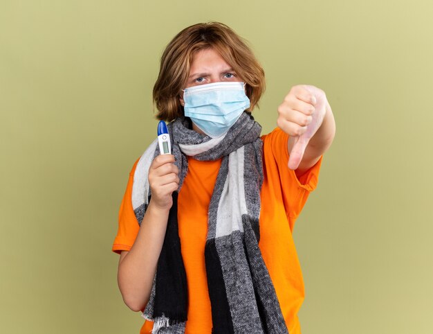 Unhealthy young woman with warm scarf around her neck wearing protective facial mask holding thermometer feeling unwell showing thumbs down