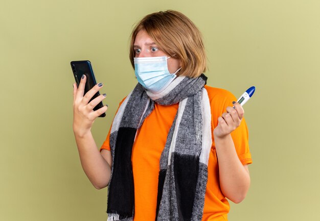 Unhealthy young woman with warm scarf around her neck wearing protective facial mask suffering from cold and flu holding thermometer talking on mobile phone looking worried on green wall