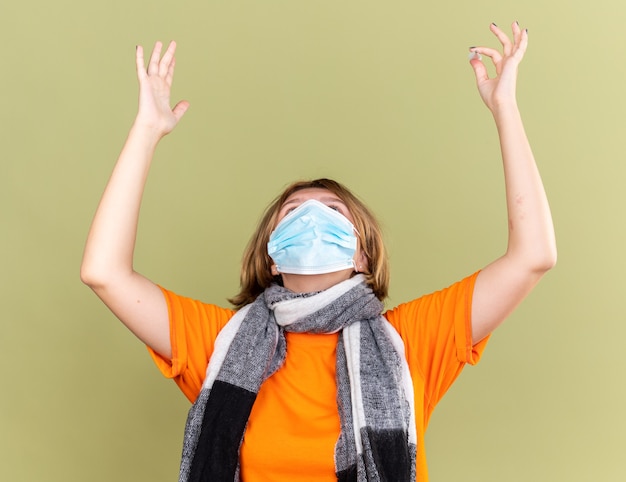 Unhealthy young woman with warm scarf around her neck wearing protective facial mask suffering from cold and flu raising hands with disappointed expression standing over green wall