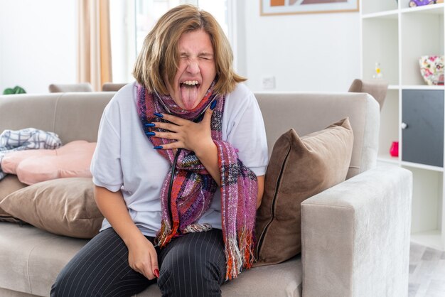 Нездоровая молодая женщина с теплым шарфом на шее чувствует себя ужасно страдающей от гриппа и простуды, прислушиваясь к своим легким с помощью стетоскопа, кашляя, находясь в панике, сидя на диване в светлой гостиной