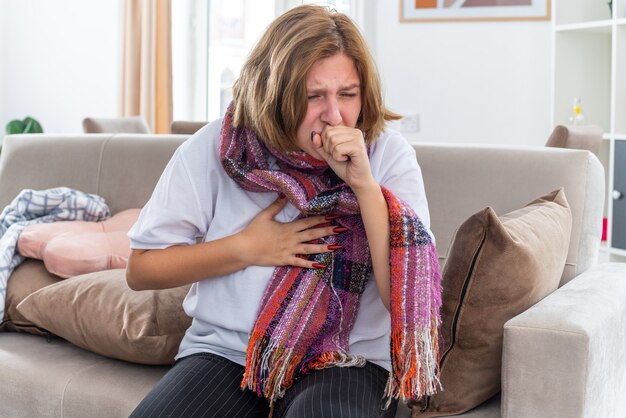 Unhealthy young woman with warm scarf around neck feeling terrible suffering from virus coughing sitting on couch in light living room