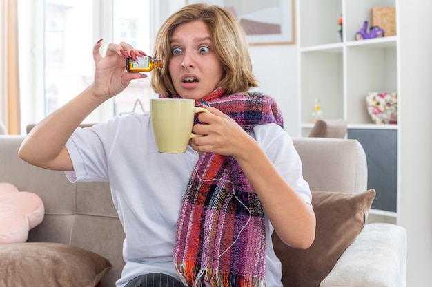 Unhealthy young woman with warm scarf around neck feeling unwell and sick suffering from flu and cold dripping medicine drops into a cup sitting on couch in light living room