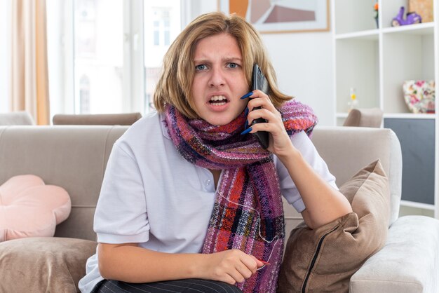 首の周りに暖かいスカーフを巻いた不健康な若い女性が、インフルエンザにかかり、風邪をひいて、明るいリビングルームのソファに座って混乱している携帯電話で話している