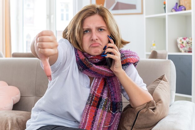 Unhealthy young woman with warm scarf around neck feeling unwell and sick suffering from flu and cold talking on mobile phone showing tumbs down sitting on couch in light living room