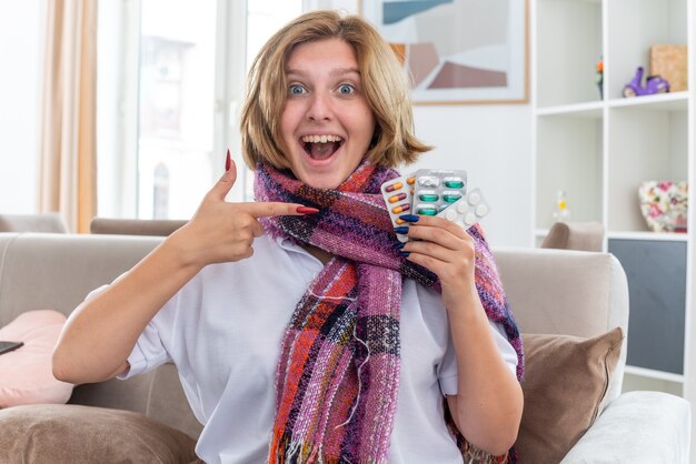 Unhealthy young woman with warm scarf around neck holding different pills pointing with index finger at them smiling cheerfully feeling better sitting on couch in light living room