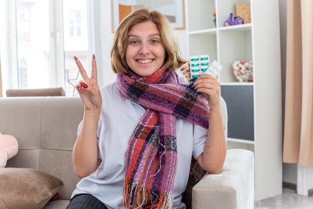 Unhealthy young woman with warm scarf around neck holding different pills  smiling showing v-sign smiling feeling better sitting on couch in light living room