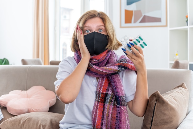 Бесплатное фото Нездоровая молодая женщина с теплым шарфом на шее с защитной маской для лица чувствует себя нездоровой и больной, страдает от гриппа и простуды, держит таблетки и выглядит смущенной, сидя на диване в светлой гостиной