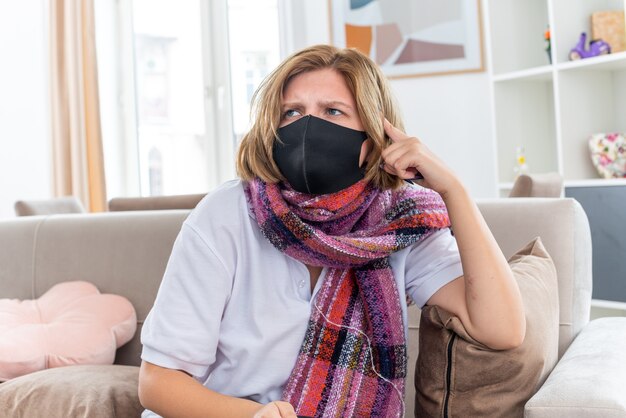 Unhealthy young woman with warm scarf around neck with facial protective mask feeling unwell and sick suffering from flu and cold looking confused sitting on couch in light living room