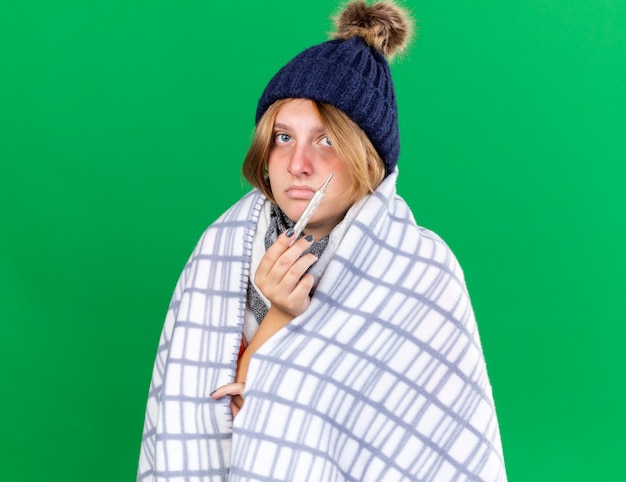Giovane donna malsana avvolta in una coperta che indossa un cappello che misura la sua temperatura corporea usando un termometro che soffre di influenza con febbre in piedi sul muro verde