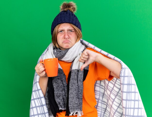 Unhealthy young woman wrapped in blanket wearing hat and scarf drinking hot tea suffering from cold feeling sick showing thumbs down standing over green wall