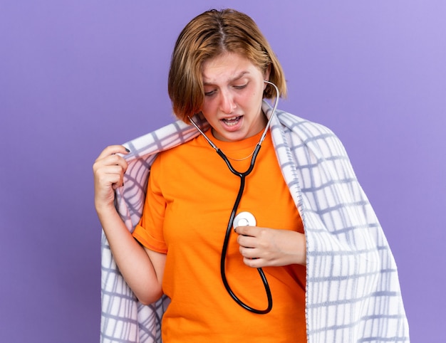 Unhealthy young woman wrapped in warm blanket feeling sick listening to her heartbeat using stethoscope looking worried standing over purple wall