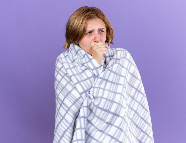 Нездоровая молодая женщина, завернутая в теплое одеяло, чувствует себя больной, страдает гриппом и кашляет с лихорадкой