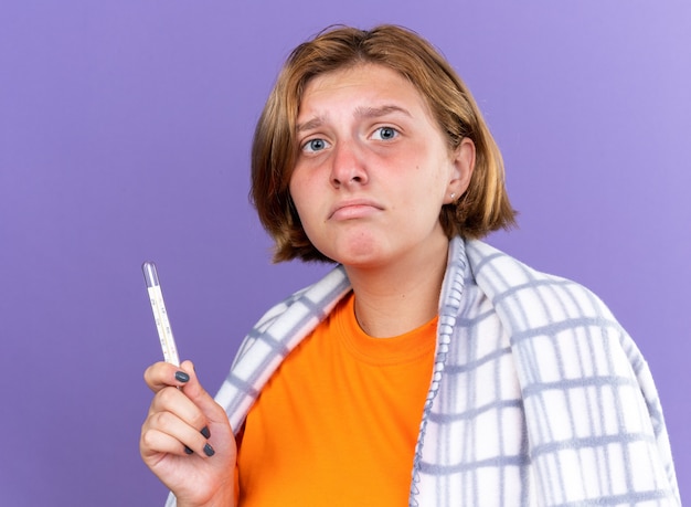 暖かい毛布に包まれた不健康な若い女性がインフルエンザにかかり、紫色の壁の上に立って心配しているように温度計を使って体温を測定している