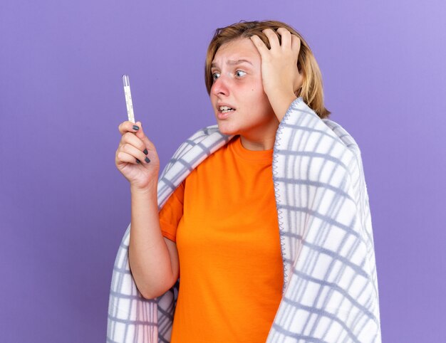 Unhealthy young woman wrapped in warm blanket feeling sick suffering from flu having fever measuring her temperature using thermometer looking worried standing over purple wall