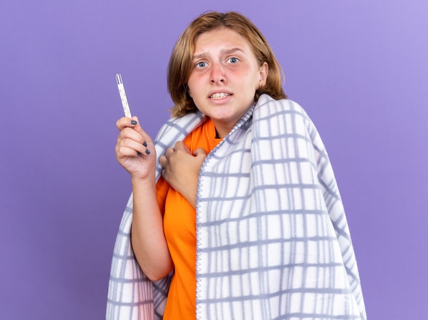 Unhealthy young woman wrapped in warm blanket feeling sick suffering from flu having fever measuring her temperature using thermometer looking worried standing over purple wall