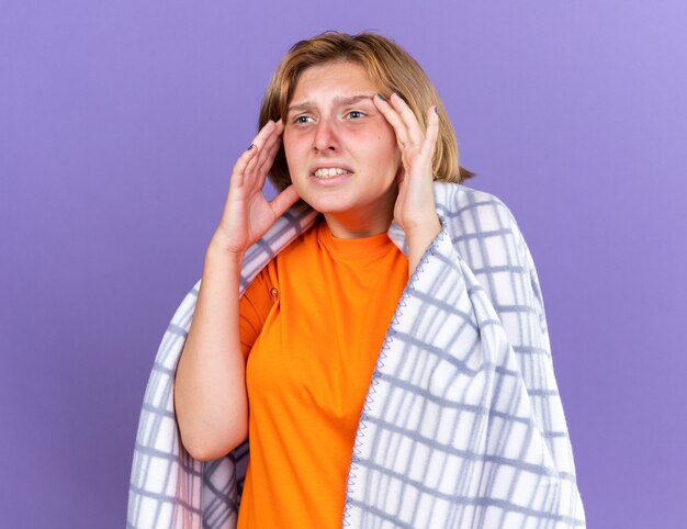 Unhealthy young woman wrapped in warm blanket suffering from flu and headache having fever measuring her temperature using thermometer looking worried