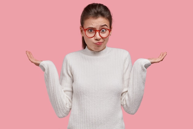 Unimpressed clueless woman spreads hands, wears glasses and white clothes, feels hesitant, makes decision in mind