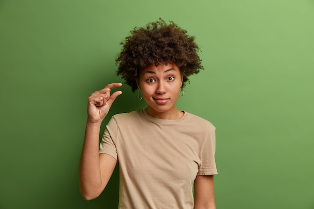 무감각 한 곱슬 여자는 약간의 제스처를 보여주고, 몇 인치 또는 센티미터, 소량 또는 낮은 규모의 기호를 보여주고, 캐주얼 한 옷을 입고 녹색 벽에 고립되어 있습니다.